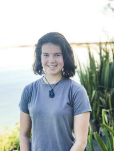 Girl in blue t-shirt standing by water with grasses behind.