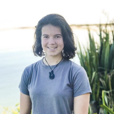 Girl in blue t-shirt standing by water with grasses behind.